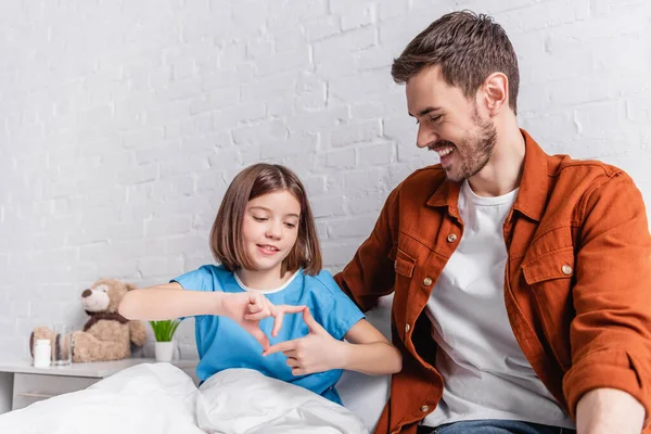 Fröhliches Mädchen gestikuliert mit Händen neben lächelndem Vater, der sie im Krankenhaus besucht — Stockfoto