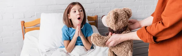 Amazed girl showing wow gesture near dad with teddy bear in hospital, banner — Stock Photo