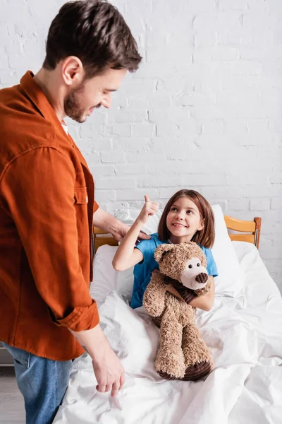 Glückliche Tochter hält Teddybär in der Hand und zeigt Vater im Krankenhaus Daumen hoch — Stockfoto