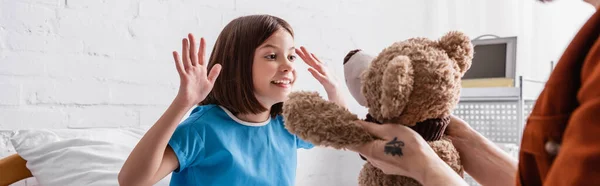 Happy child gesturing near father with teddy bear in hospital, blurred foreground, banner — Stock Photo