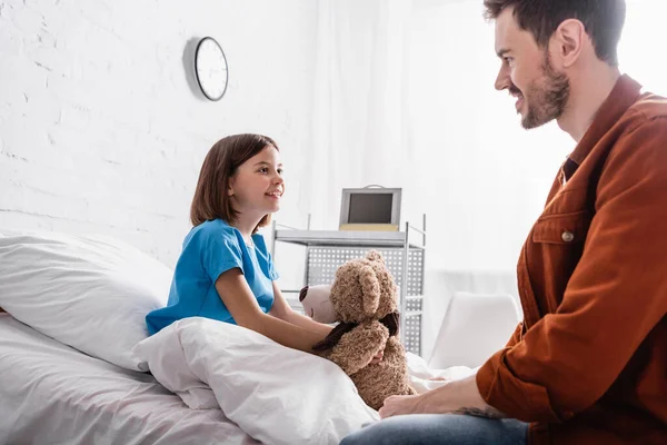 Fille souriante assise sur le lit d'hôpital avec ours en peluche près de papa heureux, avant-plan flou — Photo de stock