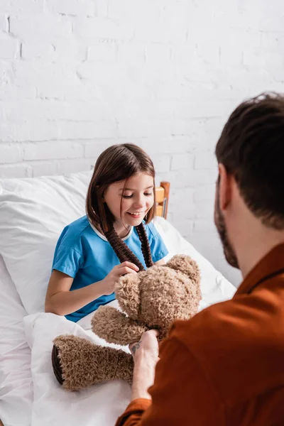 Glückliches Mädchen berührt Teddybär nahe Vater auf verschwommenem Vordergrund im Krankenhaus — Stockfoto