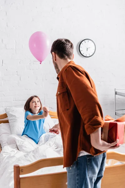 Ragazza felice tenendo palloncino festivo vicino al padre con scatola regalo in ospedale — Foto stock