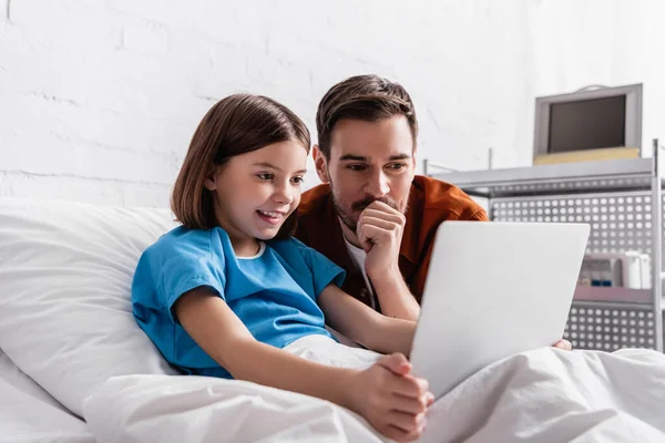Fröhliches Mädchen mit Laptop im Krankenhausbett neben glücklichem Vater — Stockfoto