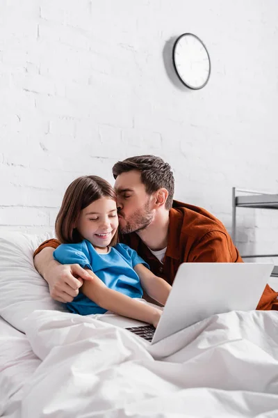 Feliz pai beijando alegre filha usando laptop na cama do hospital — Fotografia de Stock
