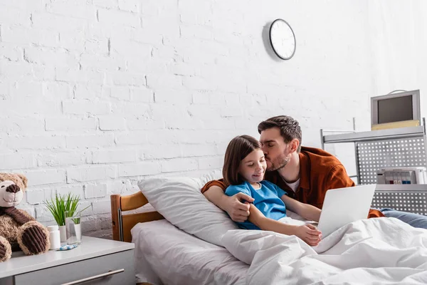 Man kissing cheerful daughter using laptop in hospital — Stock Photo