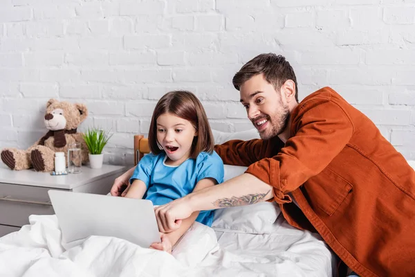 Astonished man pointing at laptop near amazed daughter in hospital bed — Stock Photo