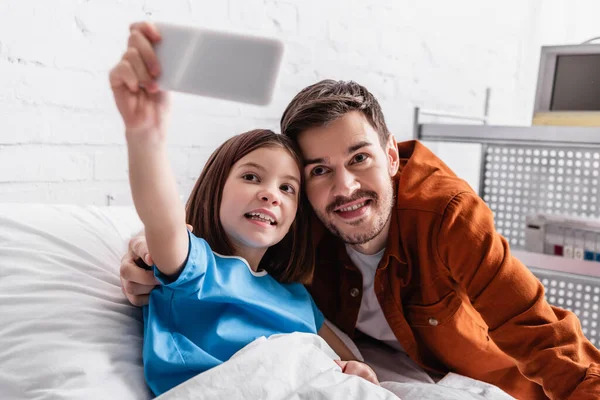 Cheerful father and daughter taking selfie on mobile phone in hospital, blurred foreground — Stock Photo