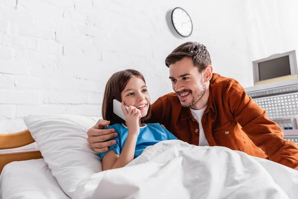 Feliz hombre abrazando sonriente hija hablando en el teléfono inteligente en cama de hospital - foto de stock