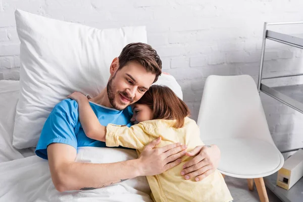 Sonriente hombre abrazando hija mientras acostado en la cama en el hospital - foto de stock