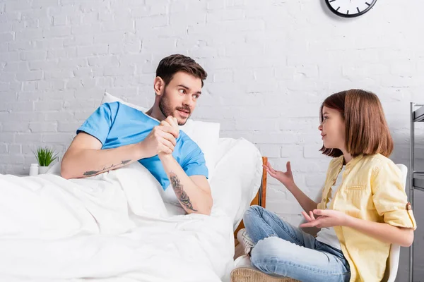 Улыбающаяся девушка жестикулирует, разговаривая с отцом, лежащим в постели в больнице — стоковое фото