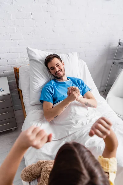 Overhead view of girl showing win gesture near smiling father lying in hospital bed, blurred foreground — Foto stock