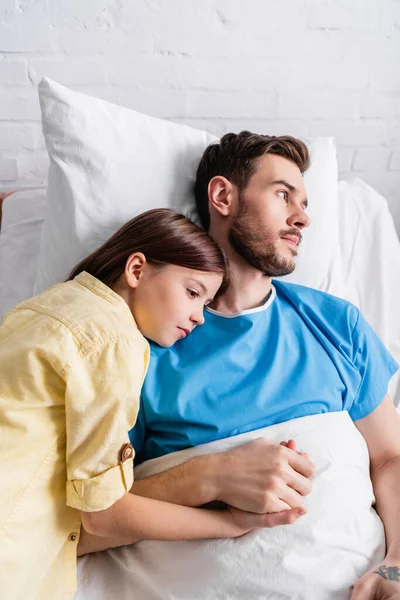 Menina inclinando-se no pai deitado na cama do hospital e segurando sua mão — Fotografia de Stock