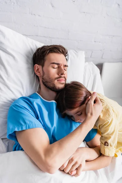 Homem doente abraçando filha enquanto deitado na cama do hospital com os olhos fechados — Fotografia de Stock