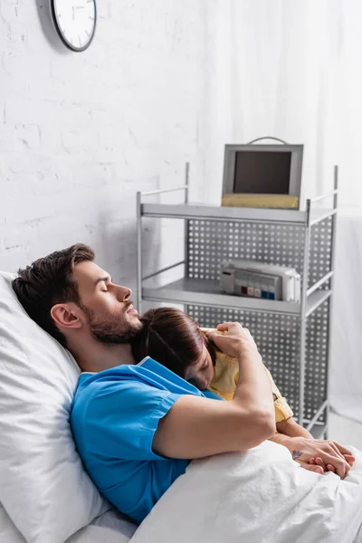 Diseased man hugging daughter while lying in bed with closed eyes — Stock Photo