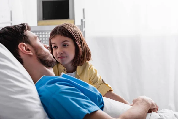 Smiling girl looking at father lying in bed in hospital, blurred foreground - foto de stock