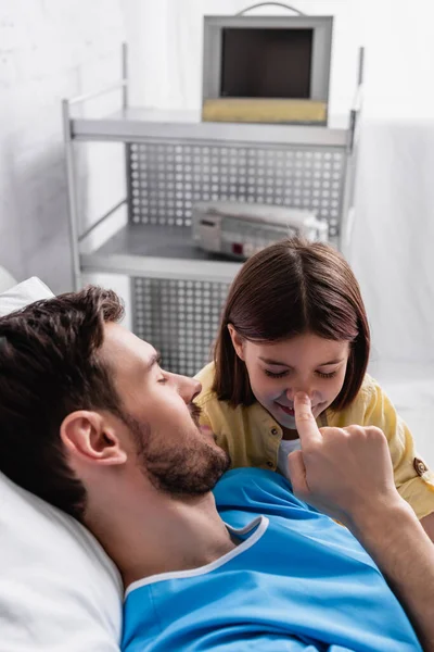 Батько торкається носа усміхненої дочки, лежачи в лікарняному ліжку — стокове фото