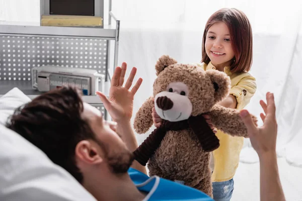 Lächelndes Mädchen gibt Teddybär an Vater, der im Krankenhaus liegt — Stockfoto