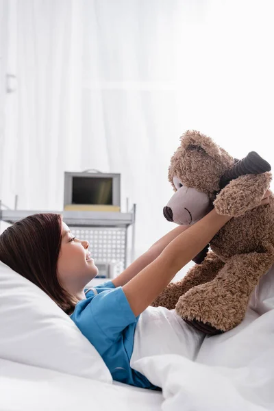 Вид збоку дитина лежить в лікарняному ліжку і тримає плюшевого ведмедя — стокове фото