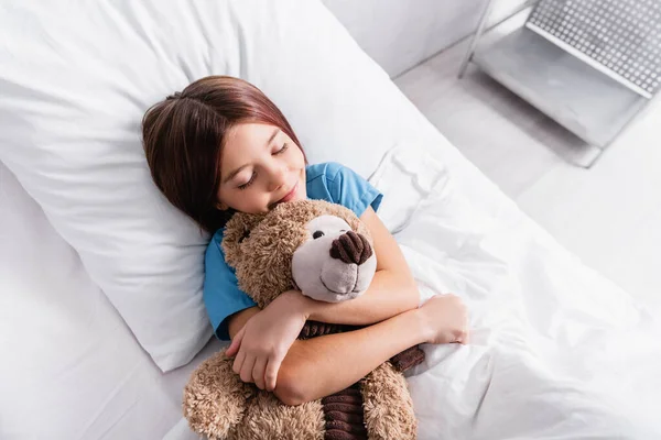 Top view of happy girl sleeping with teddy bear in hospital bed — Stock Photo