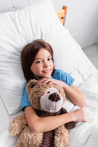 Vista superior da menina alegre olhando para a câmera enquanto deitado na cama do hospital com ursinho de pelúcia — Fotografia de Stock