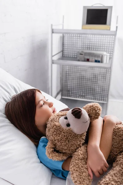 Sick, upset girl hugging teddy bear while lying in bed in hospital — Foto stock