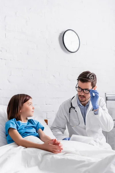 Pediatrician with digital tablet touching eyeglasses while talking to sick child in bed — Stock Photo