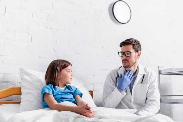 Pediatra apontando para a garganta enquanto conversa com menina na cama — Fotografia de Stock