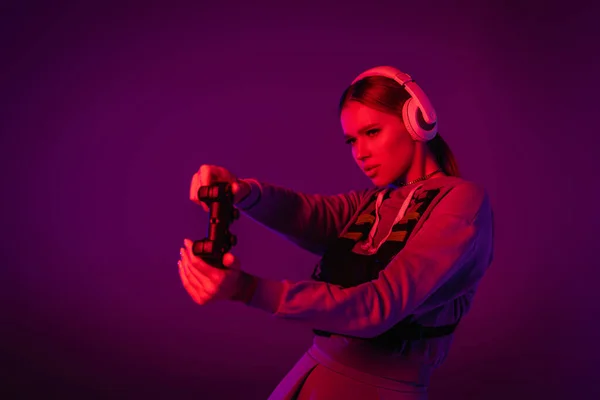 KYIV, UKRAINE - NOVEMBER 27, 2020: молода жінка в навушниках грає відеогру на фіолетовому фоні. — стокове фото