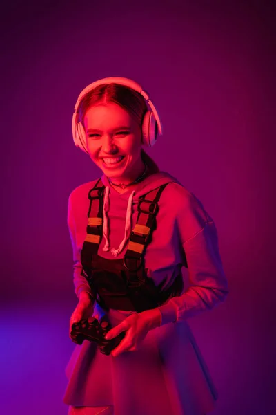 KYIV, UCRANIA - 27 de noviembre de 2020: mujer alegre en auriculares inalámbricos con joystick en púrpura - foto de stock