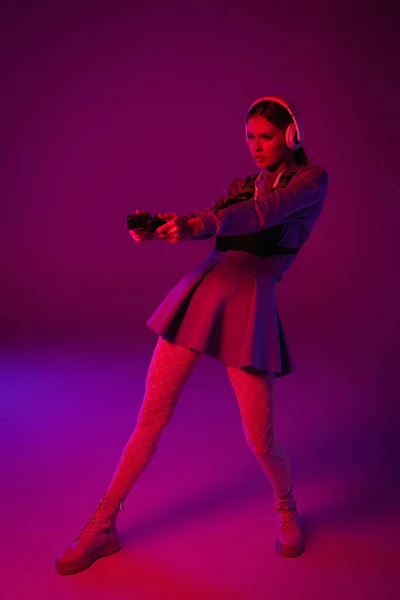 KYIV, UCRAINA - 27 NOVEMBRE 2020: tutta la lunghezza della donna in cuffie wireless con joystick su sfondo viola — Foto stock