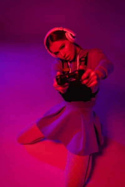 KYIV, UKRAINE - NOVEMBER 27, 2020: woman in wireless headphones holding joystick on blurred foreground and purple background — Stock Photo