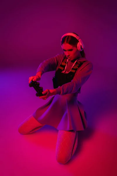 KYIV, UCRANIA - 27 de noviembre de 2020: mujer joven con auriculares inalámbricos sosteniendo el joystick y jugando videojuegos sobre fondo púrpura - foto de stock