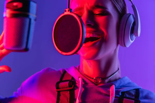 Young singer in wireless headphones recording song while singing in microphone on purple with color filter — Stock Photo