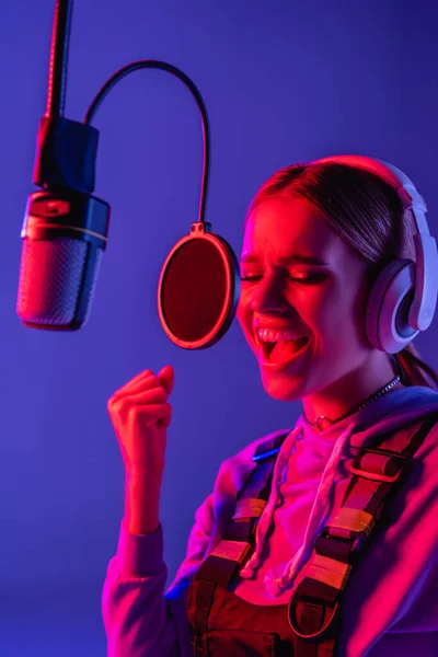 Cantante joven en auriculares inalámbricos cantando canción en micrófono en púrpura con filtro de color - foto de stock