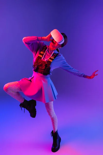 Pleine longueur de femme étonnée en réalité virtuelle casque gesticuler et sauter sur fond violet — Photo de stock