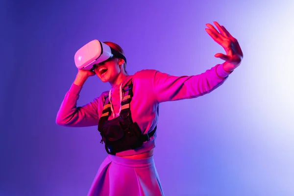 Excited young woman in virtual reality headset gesturing on purple background — Stock Photo