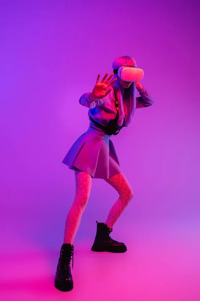 Full length of young woman in virtual reality headset standing with outstretched hand on purple background — Stock Photo