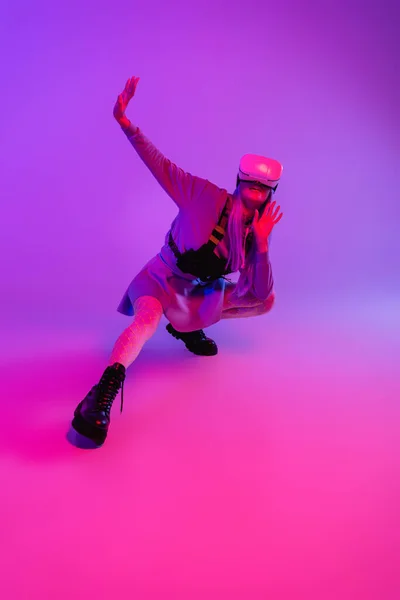 Full length of young woman in virtual reality headset sitting with outstretched hand on purple and pink — Stock Photo