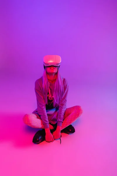 Young woman in virtual reality headset sitting with crossed legs on purple and pink — Stock Photo