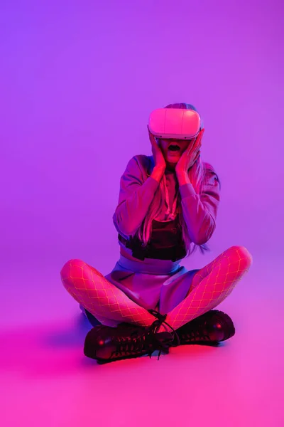 Pleine longueur de femme effrayée en réalité virtuelle casque assis sur fond violet — Photo de stock