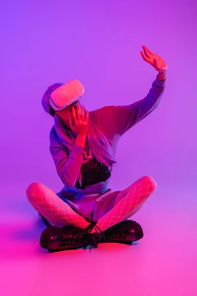 Femme surprise en réalité virtuelle casque assis avec les jambes croisées et couvrant la bouche tout en faisant un geste sur violet — Photo de stock
