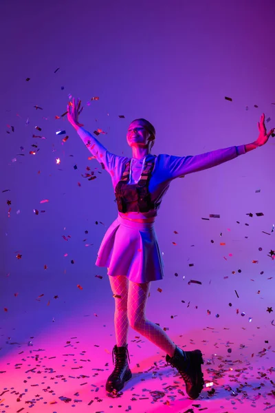 Pleine longueur de femme heureuse en tenue élégante avec les mains tendues près de confettis tombant sur violet — Photo de stock