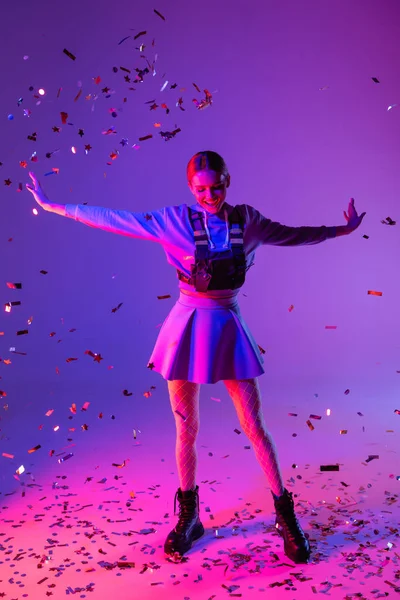 Повна довжина веселої жінки в стильному вбранні з витягнутими руками біля падаючого конфетті на фіолетовому — стокове фото
