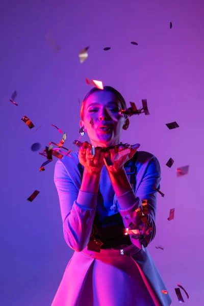 Happy woman in stylish outfit sending air kiss near falling confetti on purple — Stock Photo