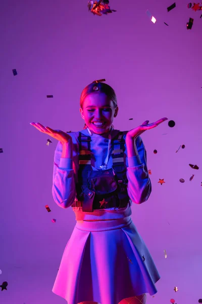Радісна жінка в стильному вбранні біля падаючого конфетті на фіолетовому — стокове фото
