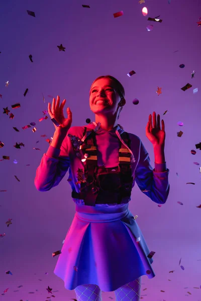 Positive woman in stylish outfit near falling confetti on purple — Stock Photo