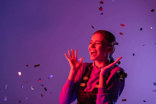 Femme gaie en tenue élégante près de confettis tombant sur violet — Photo de stock