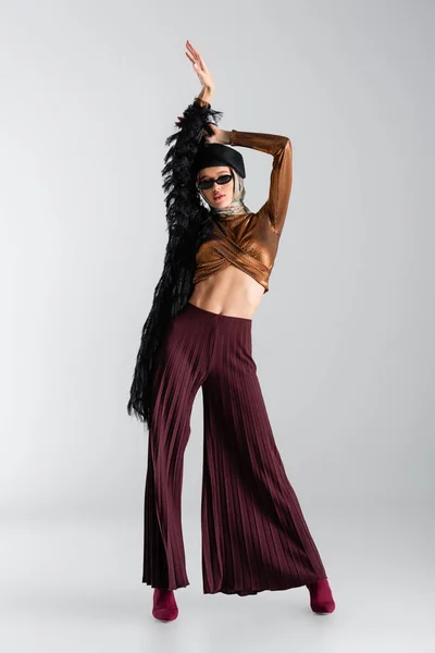 Pleine longueur de femme élégante dans des lunettes de soleil et béret posant avec les mains au-dessus de la tête sur gris — Photo de stock