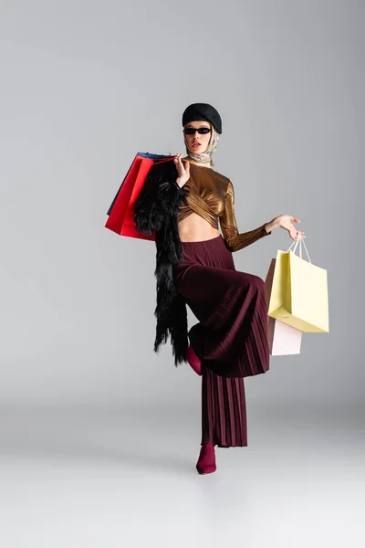 Comprimento total de mulher jovem na moda em óculos de sol e boina segurando sacos de compras e posando em cinza — Fotografia de Stock
